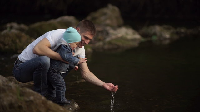 Father with little son playing on the river bank with water. Warm autumn. Daddy shows son of water in the river. Son sees dad squelching water. 