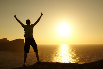 Silhouette of a man with raised arms standing on a rock on sunset. Young man traveler looking at sunset. Cap Formentor in Majorca