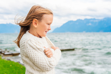 Cute little girl of 8 years old playing by the lake on a very windy day, wearing warm white knitted pullover © annanahabed