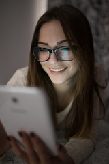 Cute woman lies on bed and using tablet in dark room. She smiling.