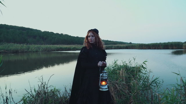young female dressed in old-fashioned clothes standing near the lake with a lamp