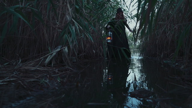 old-fashioned lady holding a lamp wading through the reed and swamp