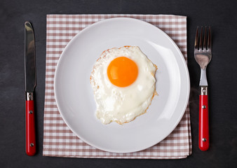 Fried egg on the plate, fork and knife on the black background.