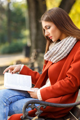 Obraz premium Young girl sitting on a park bench and reading a book, on a beautiful autumn day