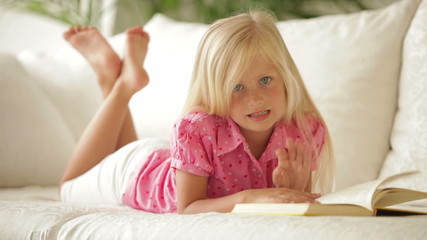 Beautiful little girl lying on sofa reading book and smiling at camera