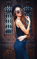 Obraz na płótnie Canvas young beautiful brunette girl in a hat, sunglasses, t-shirt and jeans, with a film camera walks in the old city, holding hand hat for fear of losing it, vertical picture