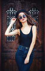 Fototapeta premium young beautiful brunette girl in a hat, sunglasses, t-shirt and jeans, with a film camera walks in the old city, holding hand hat for fear of losing it, vertical picture