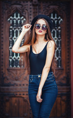 Fototapeta premium young beautiful brunette girl in a hat, sunglasses, t-shirt and jeans, with a film camera walks in the old city, holding hand hat for fear of losing it, vertical picture