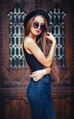 Naklejka premium young beautiful brunette girl in a hat, sunglasses, t-shirt and jeans, with a film camera walks in the old city, holding hand hat for fear of losing it, vertical picture