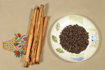 Cinnamon sticks beside a dish with black pepper grains, on an embroidered cloth