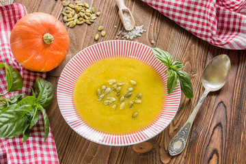 cream soup from pumpkin and other vegetables decorated by basil leaf and pumpkin seeds, basil, pumpkins fruits, salt, pumpkins  seeds  on wooden table with silver spoon on red squared  napkin