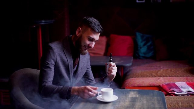 Modern hipster businessman drinking coffee in cafeand working on tablet smoking e-cigarette vape
