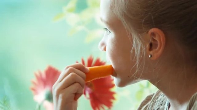 Extreme Closeup Portrait Girl Eating Carrot And Smiling At Camera. Child Sitting On A Window Sill. Child Who Loves Healthy Food. Child Looks At The Carrot