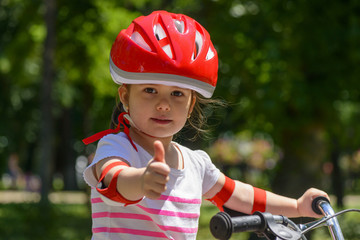 Fototapeta na wymiar Close-up of a beautiful little girl with colourful red safety helmet, showing a thumbs up, learning to ride a bike in summer park outdoors 