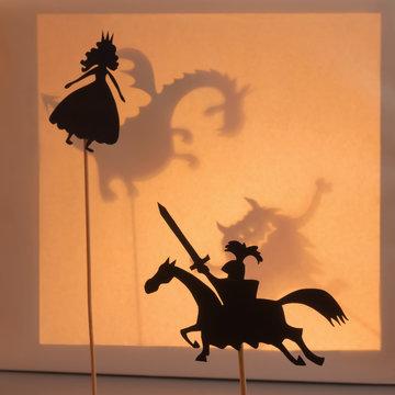 Princess and Knight shadow puppets. Bright yellow glowing screen of shadow theatre with two monsters shadows in the background. 