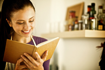 A young woman standing in the kitchen reading a cookbook.