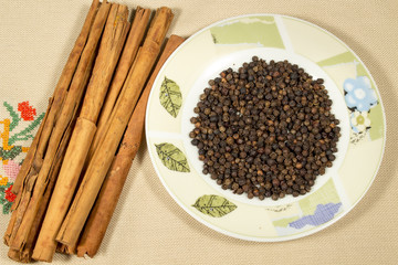 Small dish with black pepper grains beside cinnamon sticks, on an embroidered cloth