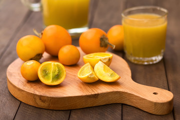 Naranjilla or Lulo fruits (lat. Solanum quitoense) on wooden board with freshly prepared naranjilla juice in the back (Selective Focus, Focus on the naranjilla pieces in the front)