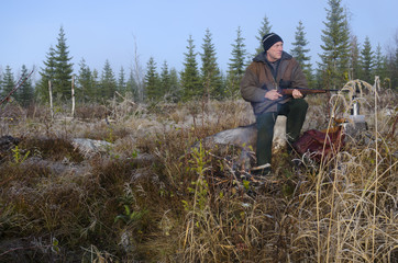 Naklejka na ściany i meble Moose hunter sitting on a stump with a litle fire in front holding his rifle pointing to the left, picture from the North of Sweden. 