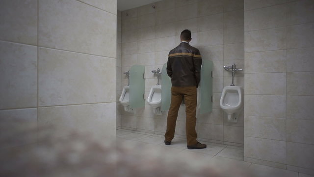Man walks into a public washroom and uses the urinal.