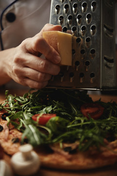 Woman cook in uniform preparing italian pizza with ham, cheddar cheese and a lot of greens in kitchen.  Chef slicing cheese on pizza. 