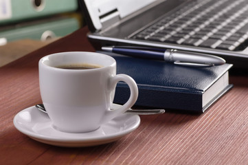 Desktop with coffee cup, opened laptop computer, diary and pen on background, no people, focused on coffee