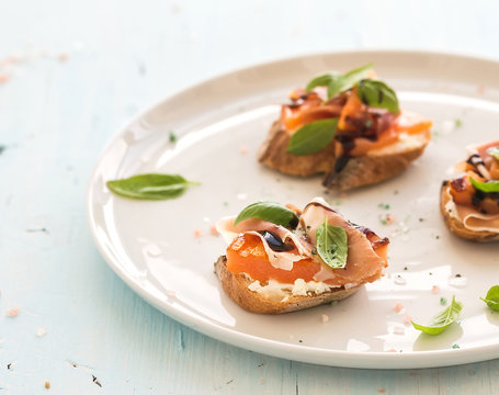 Bruschettas with Prosciutto, roasted melon, soft cheese and basil on white ceramic plate over light blue background
