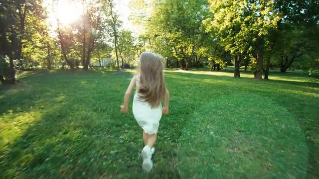 Girl running away from camera in the park. Happy child in the white dress. Lens flare
