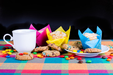Fresh homemade muffins and cookies with color chocolate coated candies, cup of tea on a colorful checkered kitchen towel. Free space for your text