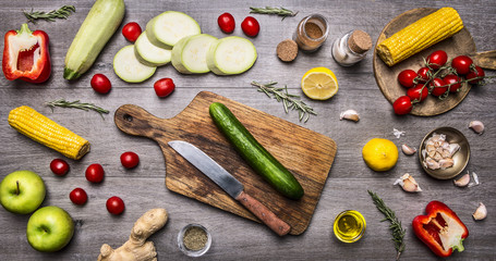 Ingredients for cooking vegetarian food peppers, corn, cucumbers, apples, cherry tomatoes, lemon, zucchini, cutting board, knife, seasoning on wooden rustic background top view