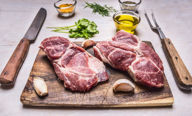 Two juicy fresh raw pork steak on a cutting board with garlic, oil, herbs, a knife and fork on wooden rustic background top view close up