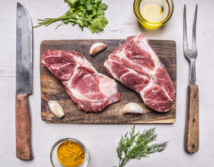 Two juicy fresh raw pork steak on a cutting board, with butter, herbs, knife and fork on wooden rustic background top view close up
