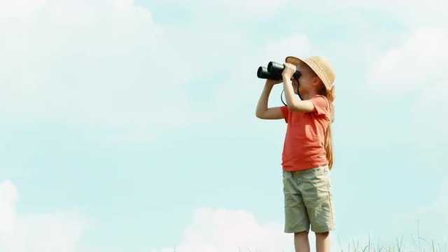 Girl through binoculars watching wildlife and looking and waving hand at camera. Girl standing against the sky