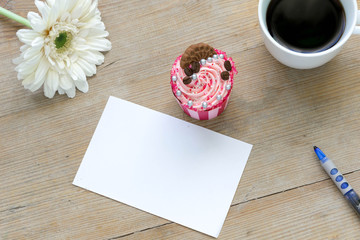 Blank paper with coffee, cupcake and flower on a wooden table, top view.