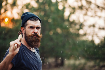 Brutal brunette bearded man in warm hat with a hatchet in the woods on a background of trees