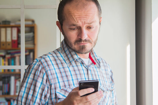 Man with beard in plaid whirt next to window typing on smart phone indoor, facial expression, communication concept