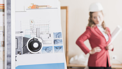 female architect in a red suit standing behind a drawing board with pen and folder in blur. Wearing glasses in the office. Planning, busy, concentration, smiling.