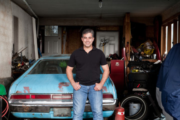 Man standing in his cluttered garage next to an old car.
