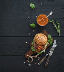 Fresh homemade burger on dark serving board with spicy tomato sauce, sea salt and herbs over black wooden background. Top view - obrazy, fototapety, plakaty