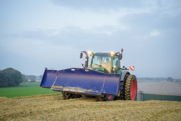 Fototapeta premium Traktor mit blauem Planierschild beim verdichten eines Maishaufens während der Dämmerung