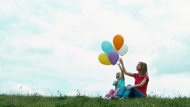 Mother and daughter with balloons sitting on the grass against the sky and playing with balloons