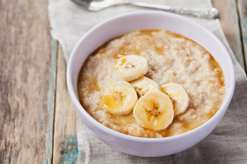 Bowl of oatmeal porridge with banana and caramel sauce on rustic table, hot and healthy breakfast every day, diet food