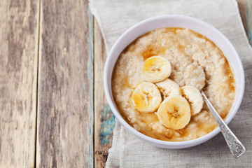 Bowl of oatmeal porridge with banana and caramel sauce on rustic table, hot and healthy breakfast every day, diet food - obrazy, fototapety, plakaty
