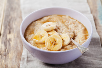 Bowl of oatmeal porridge with banana and caramel sauce on rustic table, hot and healthy breakfast...