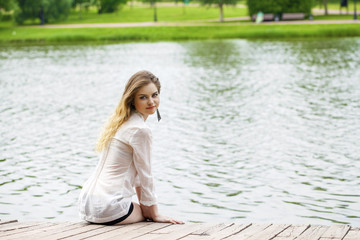 Young beautiful girl in a white tunic sits on a wooden pier on t