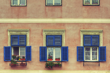 Windows with wide open blue wooden shutters and decorative blooming flowers in pots.