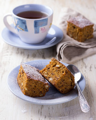 Homemade carrot and banana cake with nuts and spices, sprinkled with powdered sugar and a cup of tea on white vintage surface