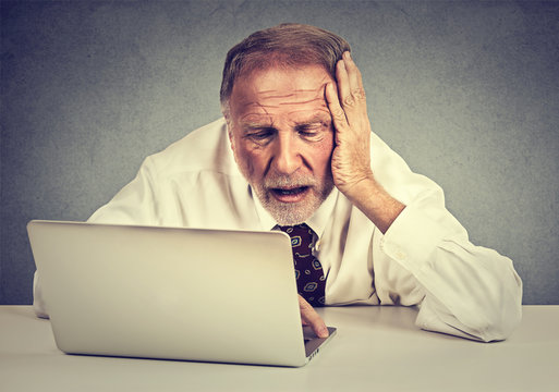 senior stressed man working on laptop sitting at table isolated on gray wall background