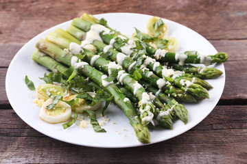 Appetizing asparagus in sour cream sauce with onion and cheese on white plate against wooden background