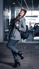 young woman with long hair, with a leather backpack, a warm hat, beautiful makeup, leather pants, a gray sweatshirt and a leather shirt, high heels walking one in the production hall and the street